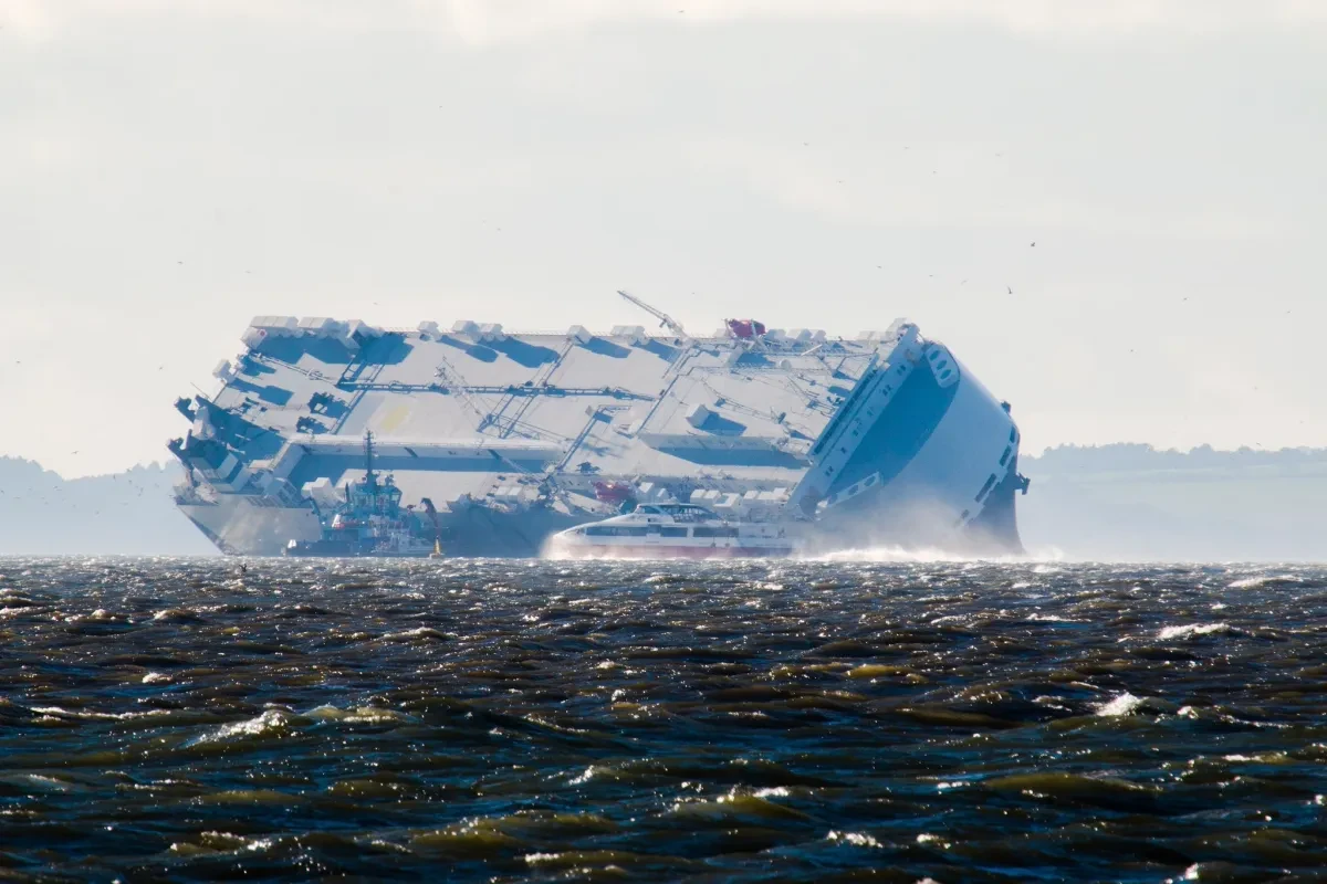 A car carrier ship on its side and stranded near the coast, with a choppy sea and a small ferry in the foreground
