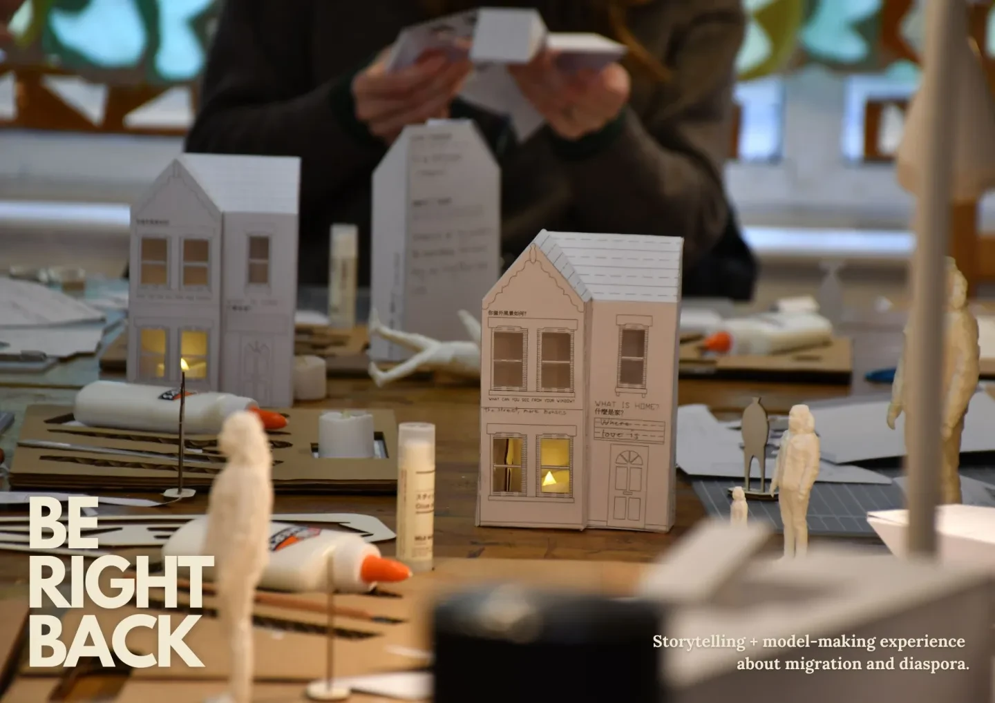 photo of paper model houses with some paper figures, in the background are the hands of a person making a paper house