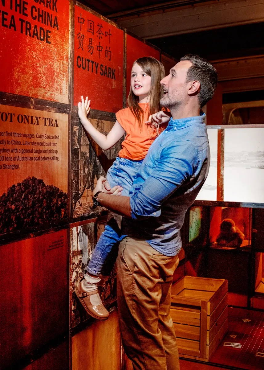 A father lifts his daughter up to look at a display of tea chests inside Cutty Sark