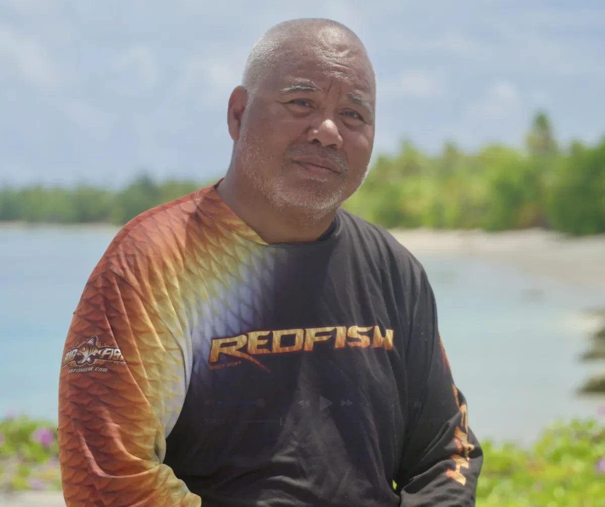Image of Alson Kelen standing on a beach wearing a black and orange t-shirt