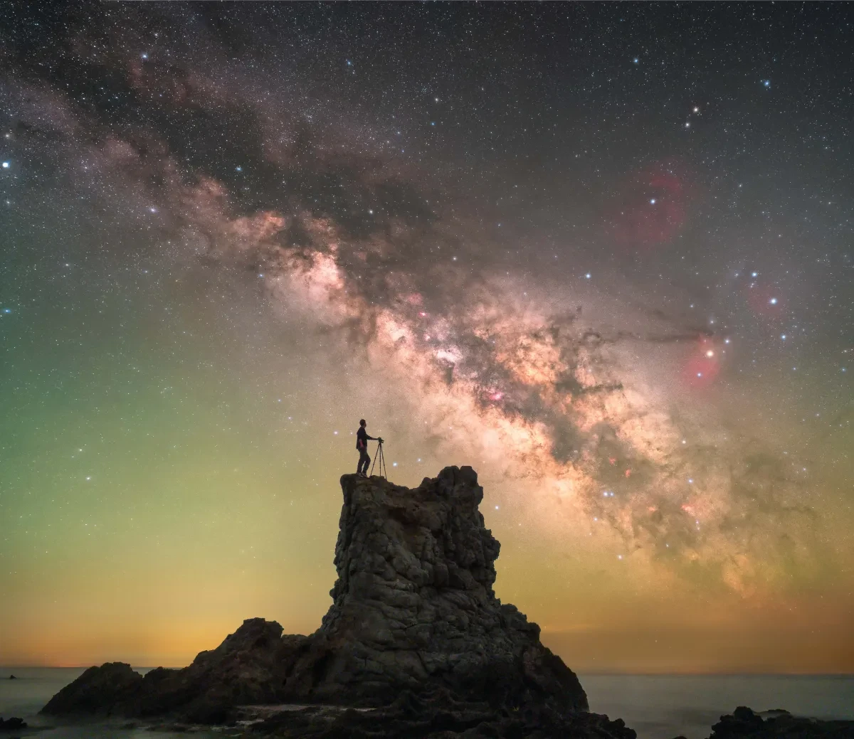 Silhouette of person on rocky outcrop with curve of Milky Way in green and yellow-tinged sky