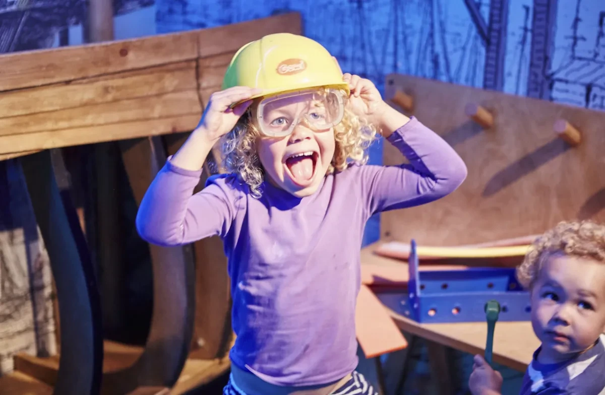 A child sticks their tongue out while wearing toy safety goggles and hard hart.