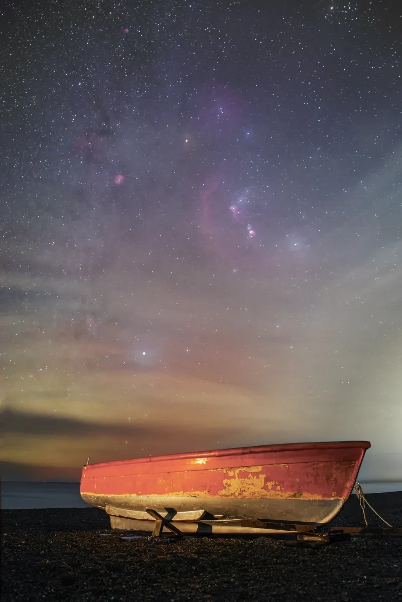 Photograph of the orange rusted hull of a small boat on a pebbly beach with the sea in the background. In the sky is a starry sky with faint purple and pink nebulae