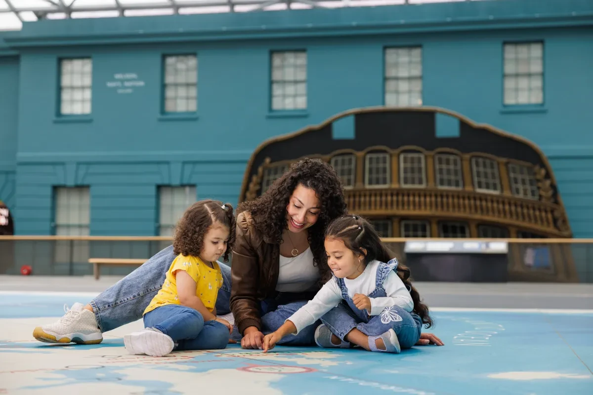 A mother and her two young daughters sit together in the centre of the National Maritime Museum. Behind them the stern of a large ship is displayed against a blue wall