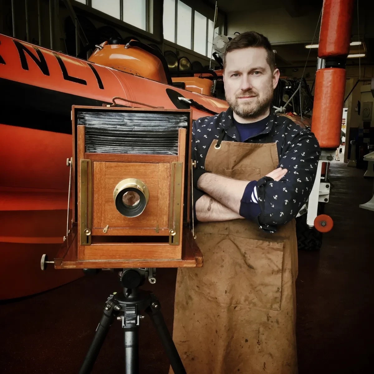 Picture of a man wearing a brown apron standing next to an Edwardian camera in front of an RNLI vessel