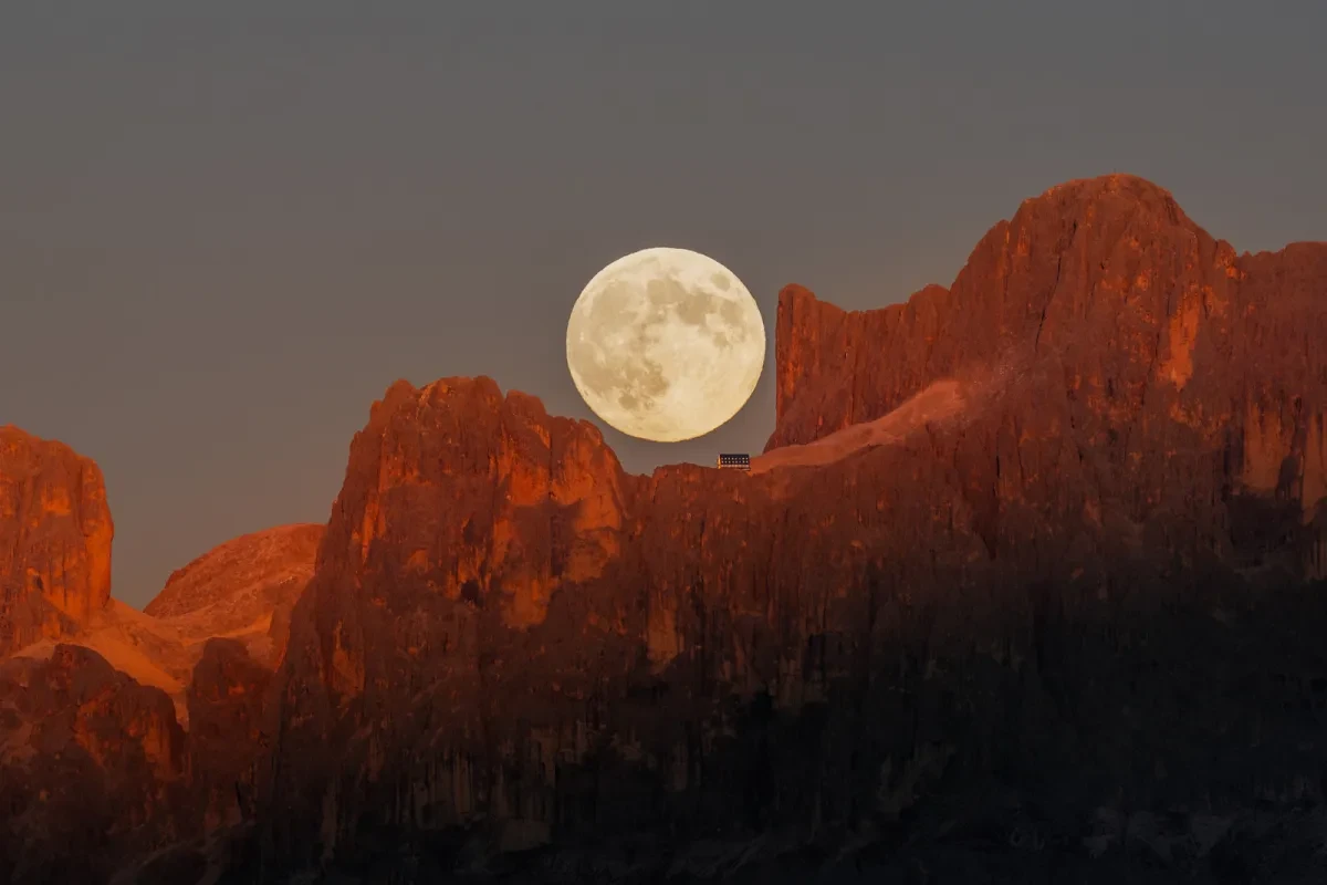 The Full Moon in a dark grey sky above red mountain peaks.