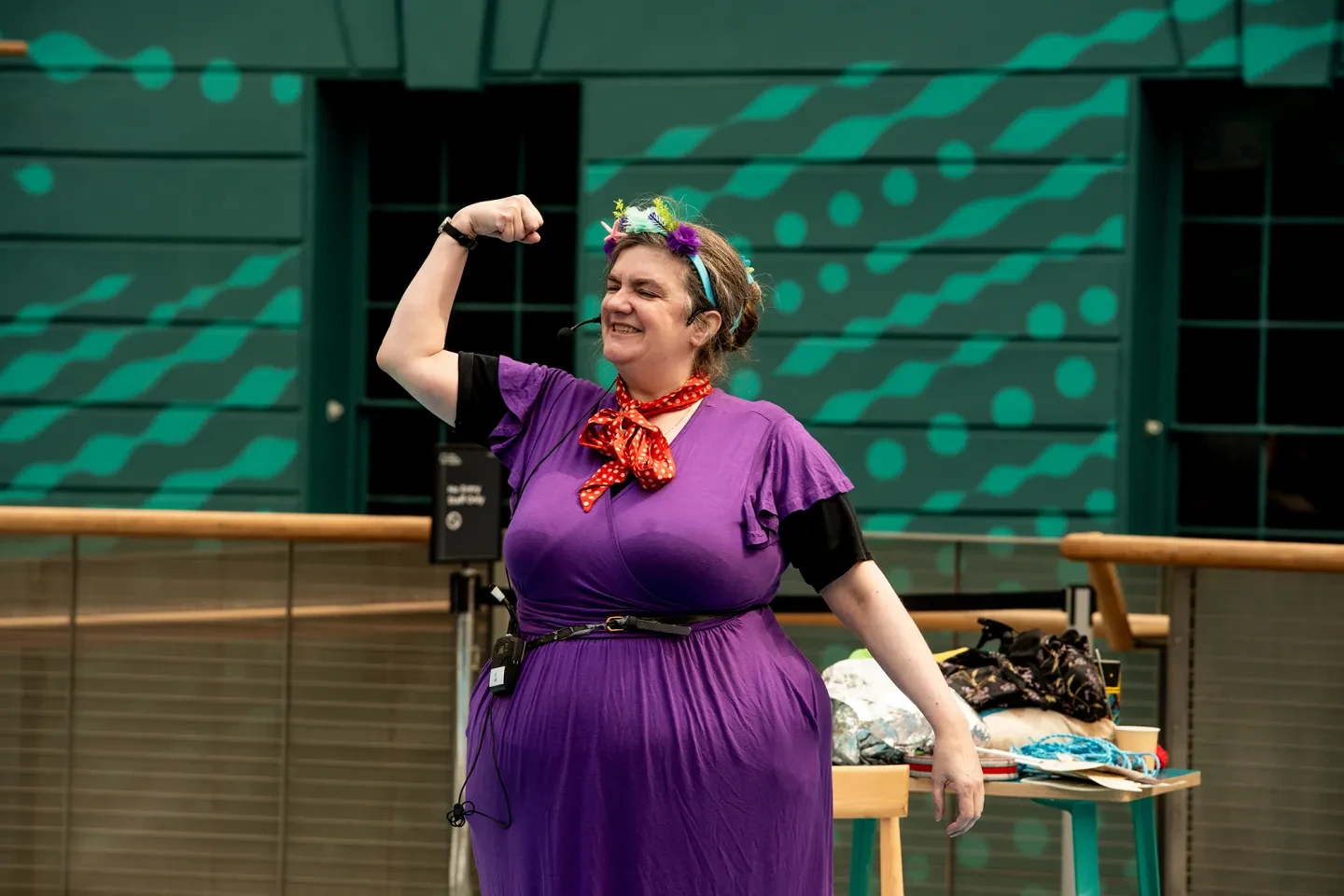 A woman in a purple dress and floral head garland stands with her arm raised and muscle flexed during a performance