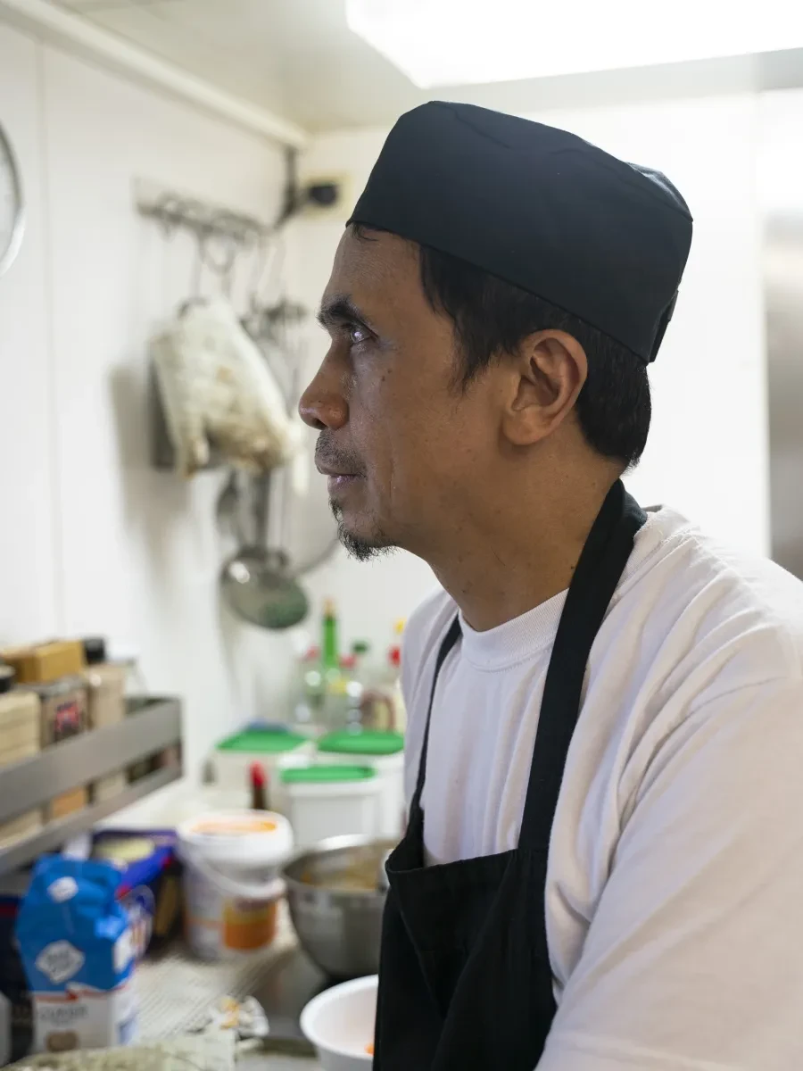 Ship's cook Reynaldo looking to one side. He is dressed in a black apron, chef's cap and white top
