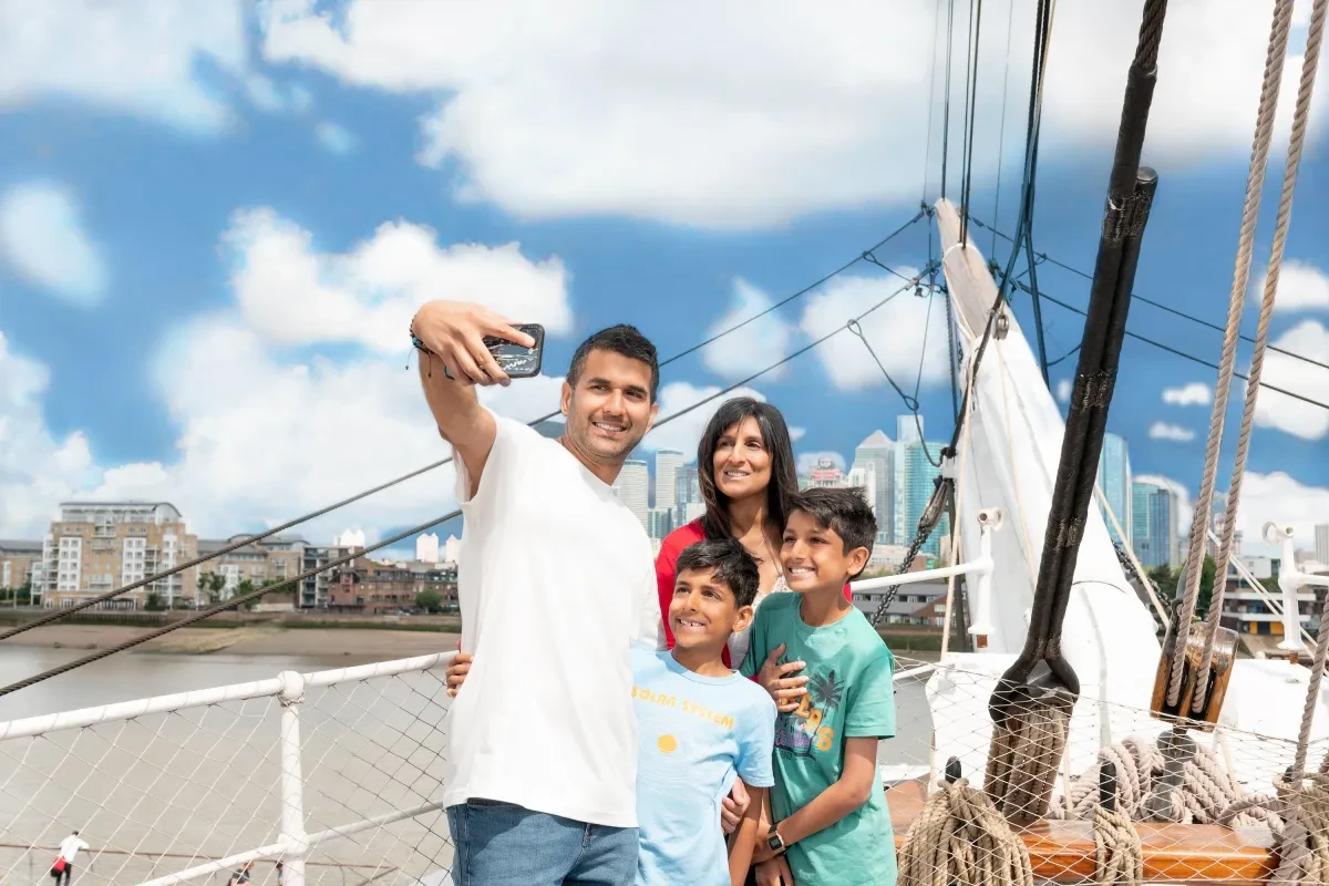 A family takes a selfie on the main deck of historic ship Cutty Sark, with Canary Wharf in the background