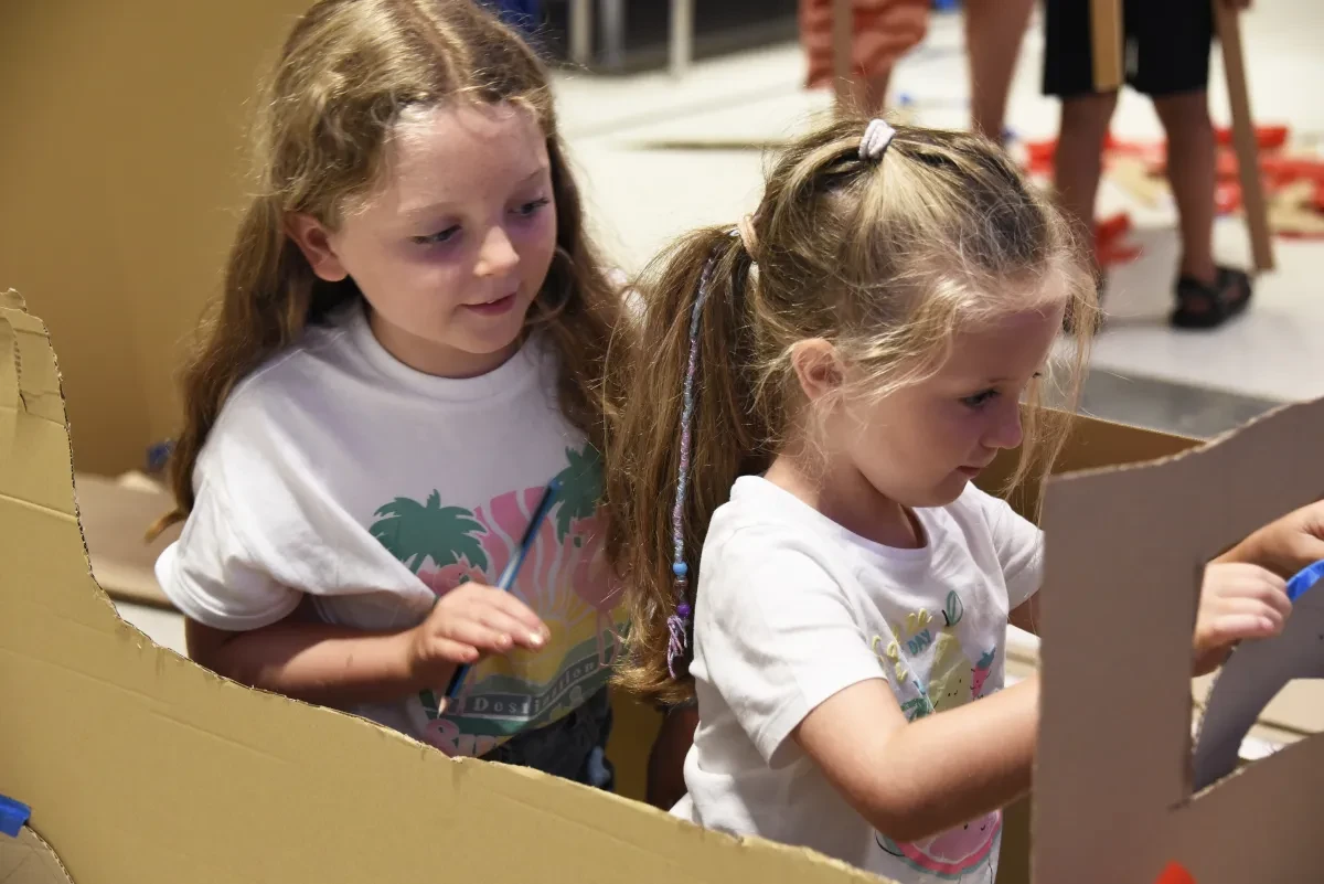 Two young girls play in a cardboard box that they have cut out into a vehicle shape