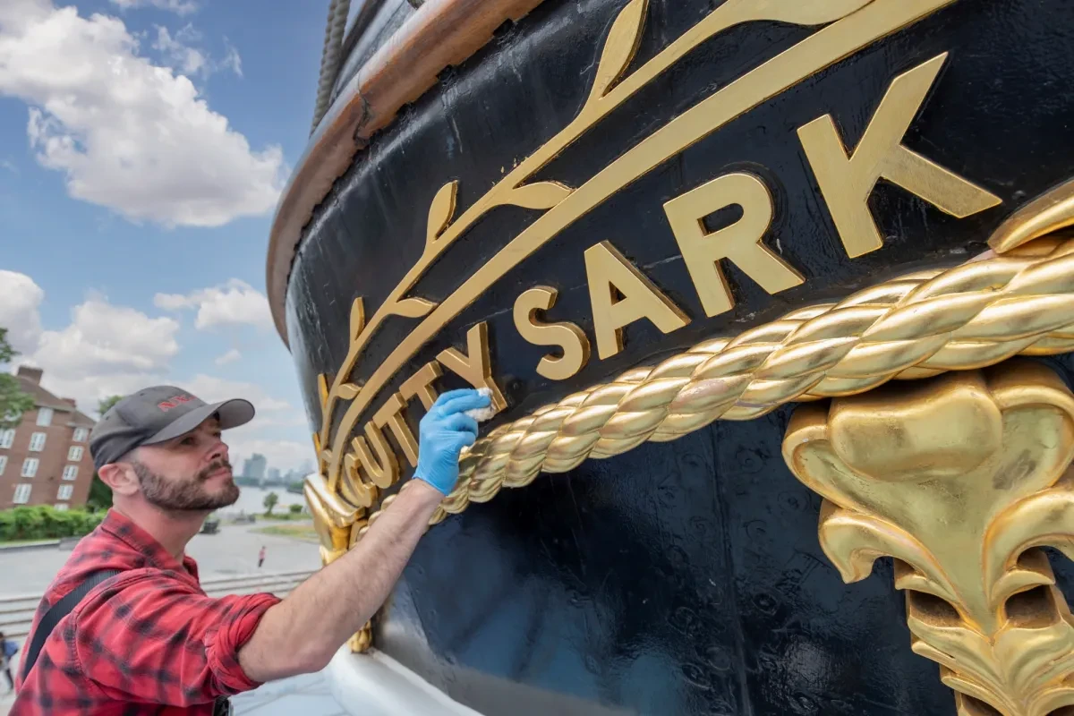 A man polishes the gold lettering of 'Cutty Sark', where the ship's name is displayed on the stern