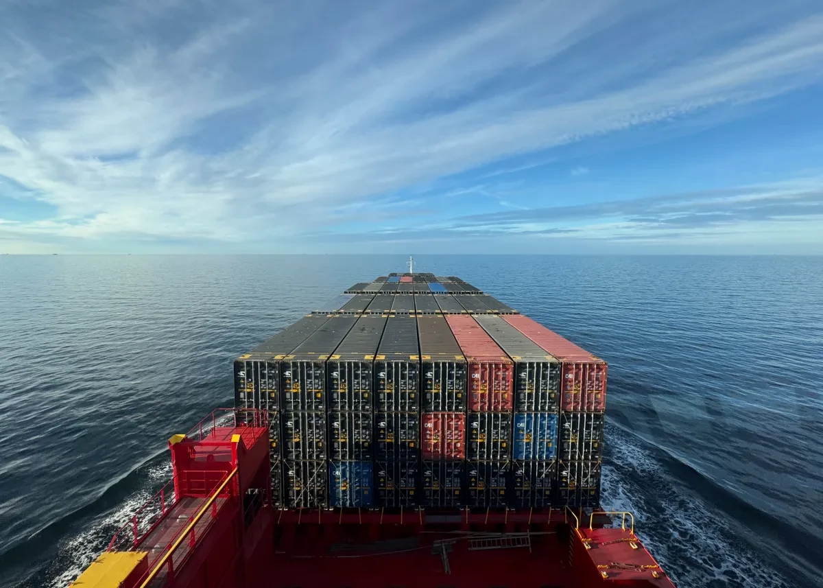 View from the bridge of a container ship at sea. A large number of containers are stacked on the deck. The sea is calm and the sky a hazy blue