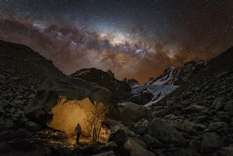 An explorer is silhouetted against a cave in front of a starry sky.